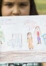 A young girl at a Peace Education camp holds up a drawing of people with speech bubbles and Russian text